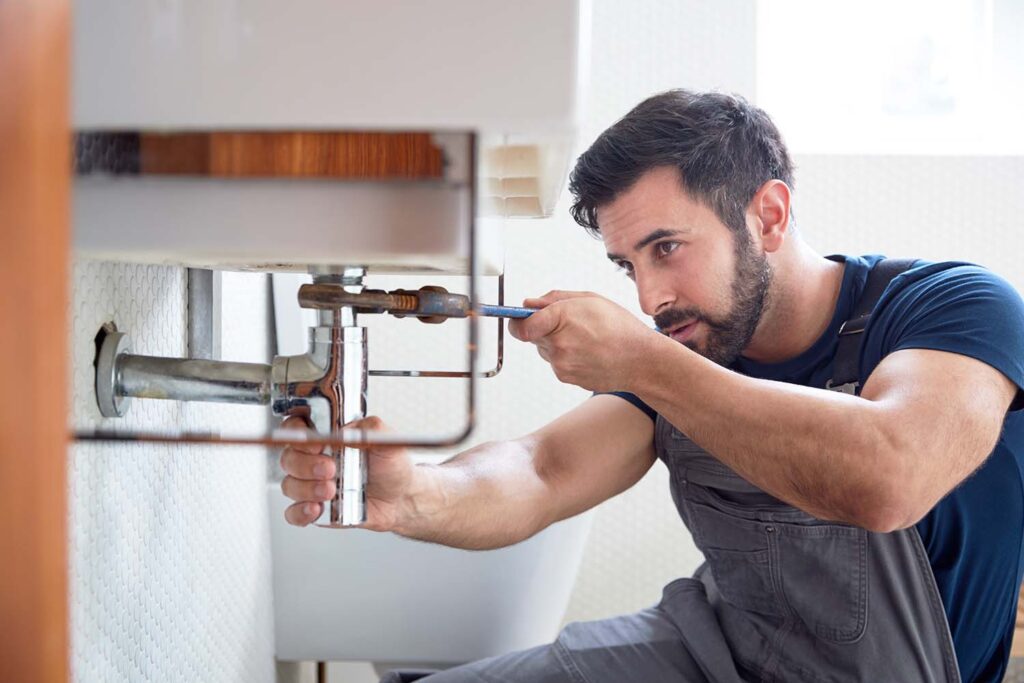 Male Plumber Using Wrench To Fix Leaking Sink In Home Bathroom